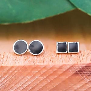 Puede incluir: Dos pares de pendientes de botón sobre una bandeja de madera. Un par es redondo con centro negro y borde plateado. El otro par es cuadrado con centro negro y borde plateado. Hojas verdes en el fondo.