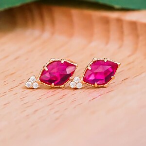 Puede incluir: Un par de pendientes dorados con grandes centros de piedras preciosas rosas vibrantes. Cada pendiente presenta un grupo de pequeñas piedras transparentes en la base. Los pendientes se exhiben en una bandeja de madera clara, con hojas verdes en el fondo.