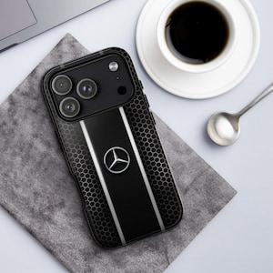 May include: Black phone case with a honeycomb pattern and silver stripes. The case features a Mercedes-Benz logo in the center. The phone case is on a gray surface next to a cup of coffee and a spoon.