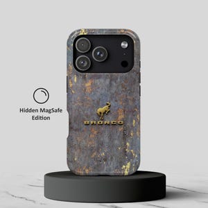 Puede incluir: Una funda para smartphone con una textura metálica desgastada en gris oscuro y color óxido. Centrado hay un emblema de caballo Bronco dorado sobre la palabra BRONCO en letras doradas. A la izquierda, el texto dice Hidden MagSafe Edition.