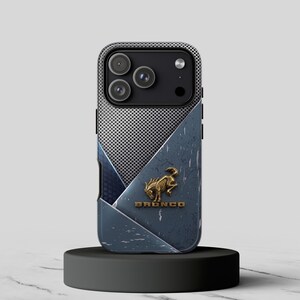 Puede incluir: Una funda para smartphone con una sección superior plateada perforada y una sección inferior azul desgastada. Un emblema dorado de un caballo encabritado está centrado sobre el texto dorado en relieve que dice BRONCO en el panel inferior.