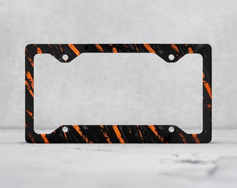 May include: A rectangular license plate frame featuring a black base with diagonal orange and grey brushstroke patterns. The frame includes four circular mounting holes positioned at the corners for attachment to a vehicle.