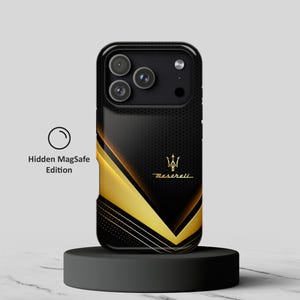 Puede incluir: Una funda para smartphone negra y dorada con textura perforada, un logotipo de Maserati dorado y un diseño gráfico en forma de V. La funda incluye un recorte para la cámara y el texto Hidden MagSafe Edition junto a un icono circular.