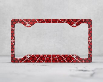 May include: A rectangular license plate frame featuring a red and black honeycomb pattern overlaid with a white web design. The frame has four mounting holes, one in each corner, and a smooth, flat surface with a consistent geometric graphic.