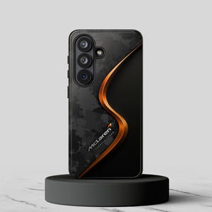 Puede incluir: Una funda negra para smartphone con un patrón de camuflaje y un diseño curvo de color cobre. El logotipo de McLaren Formula 1 Team es visible en la funda.