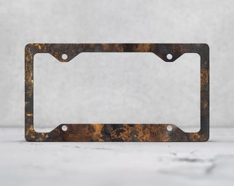 May include: A rectangular license plate frame featuring a distressed, weathered finish with mottled dark brown, black, and copper-toned metallic textures. The frame includes four circular mounting holes positioned at the corners.