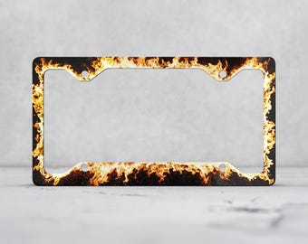 May include: A rectangular license plate frame featuring a vibrant, high-contrast graphic print of orange and yellow flames against a solid black background. The frame includes four circular mounting holes positioned at the corners for attachment.