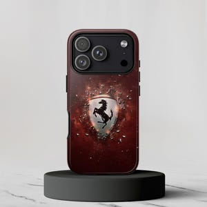 以下が含まれることがあります： フェラーリのロゴが入ったマルーン色の携帯電話ケース。ケースは、テクスチャ効果のある濃い赤色の背景と、黒い馬の入ったシルバーのエンブレムが付いています。携帯電話ケースは黒いスタンドの上にあります。