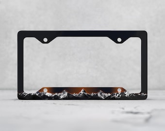 May include: A rectangular black license plate frame featuring a graphic print of snow-capped mountain peaks along the bottom edge, set against a dark sky with a subtle orange glow behind the mountain range, including four mounting holes.