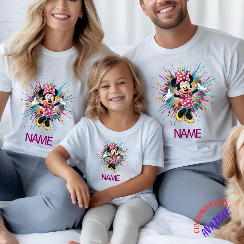 May include: White t-shirts with a Minnie Mouse graphic and a colourful burst design. The word "NAME" is printed below the graphic. The shirts are worn by three people. The shirts are part of a family matching set.