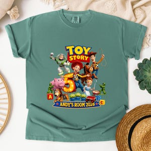 May include: A sage green short-sleeve t-shirt featuring a graphic print of Toy Story characters including Buzz Lightyear, Woody, Jessie, Bullseye, and others surrounding a large number 5. The text reads Toy Story and Andy's Room 2026.
