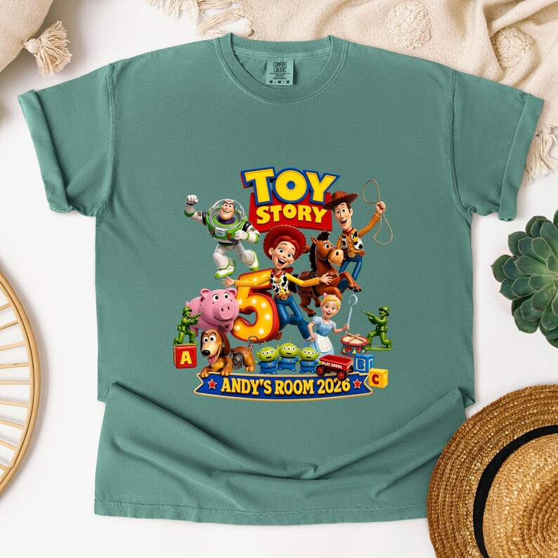 May include: A sage green short-sleeve t-shirt featuring a graphic print of Toy Story characters including Buzz Lightyear, Woody, Jessie, Bullseye, and others surrounding a large number 5. The text reads Toy Story and Andy's Room 2026.