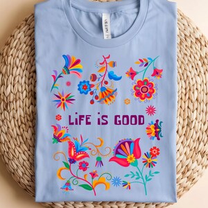 May include: A light blue t-shirt with a colorful floral design and the words "LIFE IS GOOD" in purple. The flowers are in shades of red, orange, yellow, blue, and pink. The shirt is folded on a woven mat.
