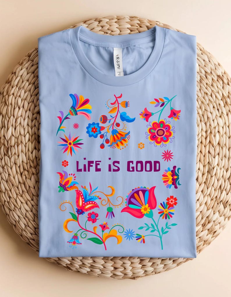May include: A light blue t-shirt with a colorful floral design and the words "LIFE IS GOOD" in purple. The flowers are in shades of red, orange, yellow, blue, and pink. The shirt is folded on a woven mat.