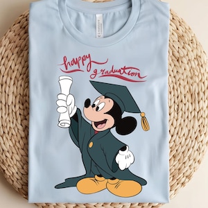 May include: Light blue t-shirt featuring Mickey Mouse in a graduation cap and gown, holding a diploma. The words "happy graduation" are written in red script above the character. The shirt is folded on a woven surface.