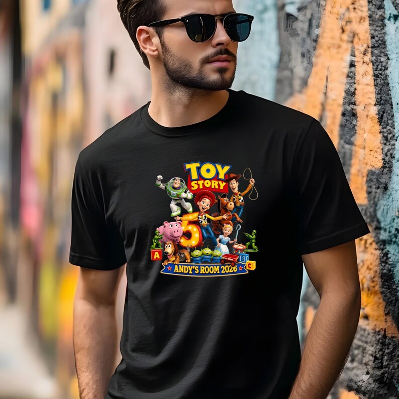 May include: A black short-sleeved t-shirt featuring a colorful Toy Story graphic print. The design includes characters like Woody, Buzz Lightyear, Jessie, and others surrounding a large number 5, with the text "TOY STORY" and "ANDY'S ROOM 2026".