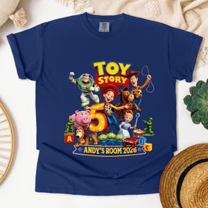 May include: A navy blue short-sleeve t-shirt featuring a colorful graphic of Toy Story characters including Woody, Buzz Lightyear, Jessie, and Bullseye. The design includes a large number 5, the text TOY STORY, and ANDY'S ROOM 2026.