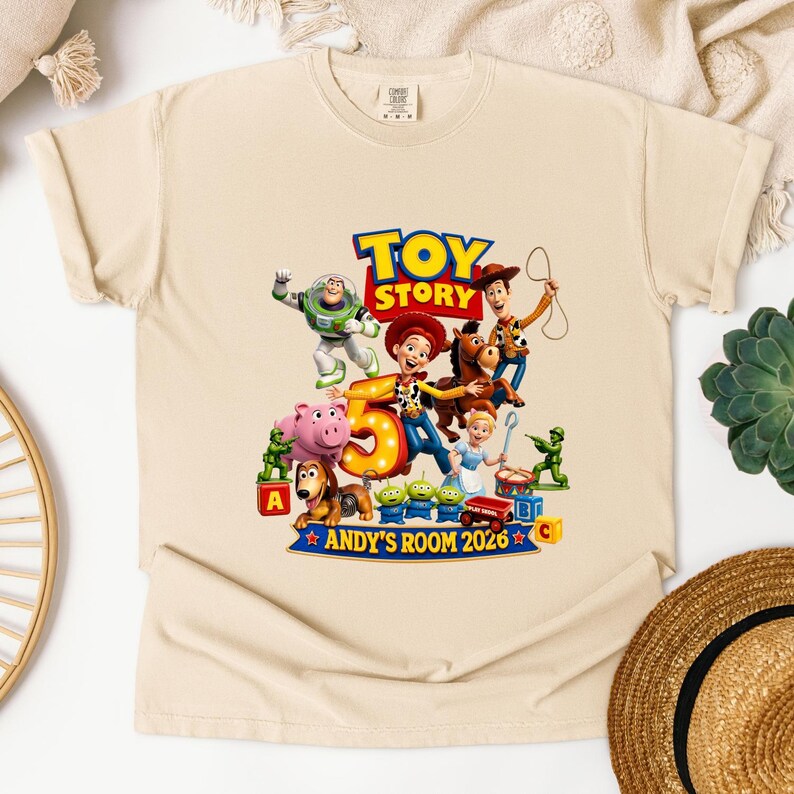 May include: A beige short-sleeve t-shirt featuring a colorful graphic of Toy Story characters including Woody, Buzz Lightyear, Jessie, and others surrounding a large number 5. The text reads TOY STORY, ANDY'S ROOM 2026, and includes alphabet blocks.