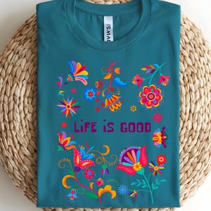 May include: Teal t-shirt with a colorful floral design and the words "LIFE IS GOOD" in a purple font. The floral design includes red, yellow, blue, and orange flowers and leaves. The shirt is displayed on a woven mat.