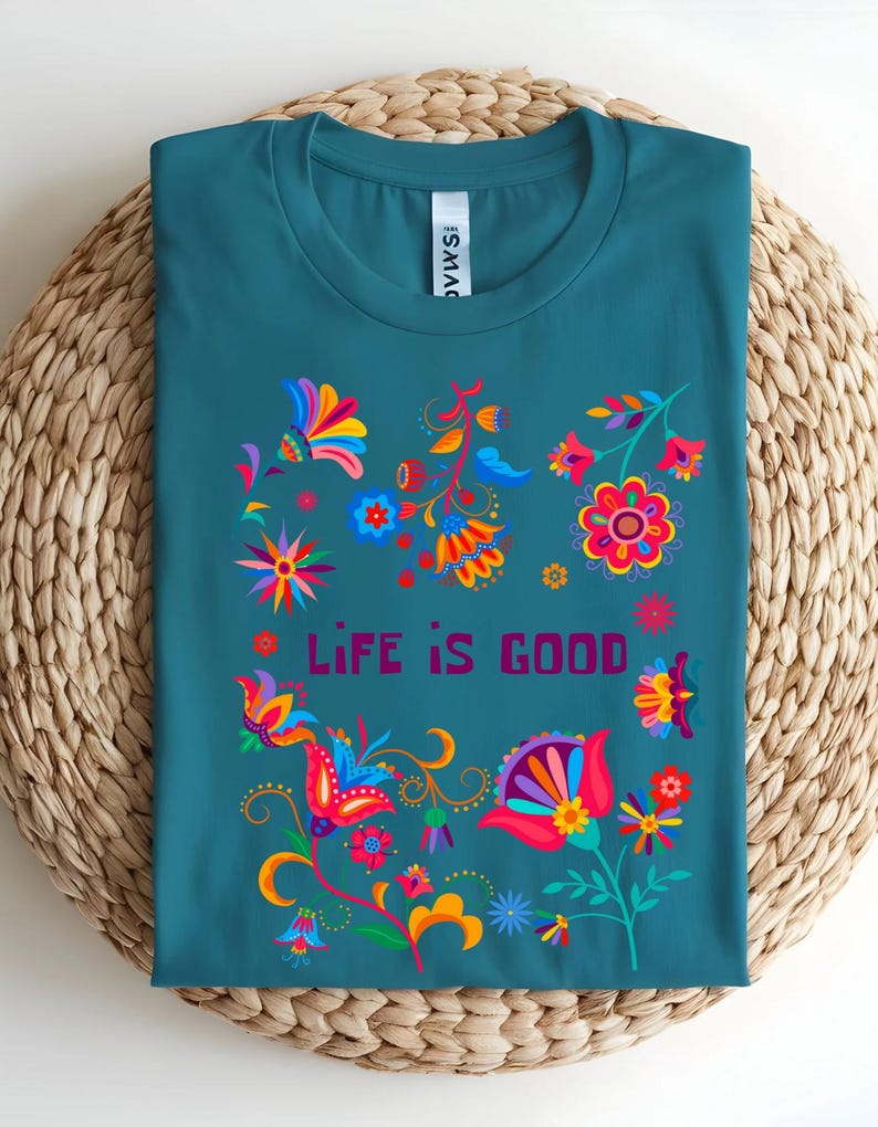 May include: Teal t-shirt with a colorful floral design and the words "LIFE IS GOOD" in a purple font. The floral design includes red, yellow, blue, and orange flowers and leaves. The shirt is displayed on a woven mat.