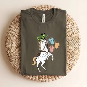 May include: Olive green t-shirt featuring a white horse rearing up, with a green chameleon and colorful balloons. The horse has a long white mane and tail. The balloons are in blue, yellow, and pink. The shirt has a tag that says "EVAS".