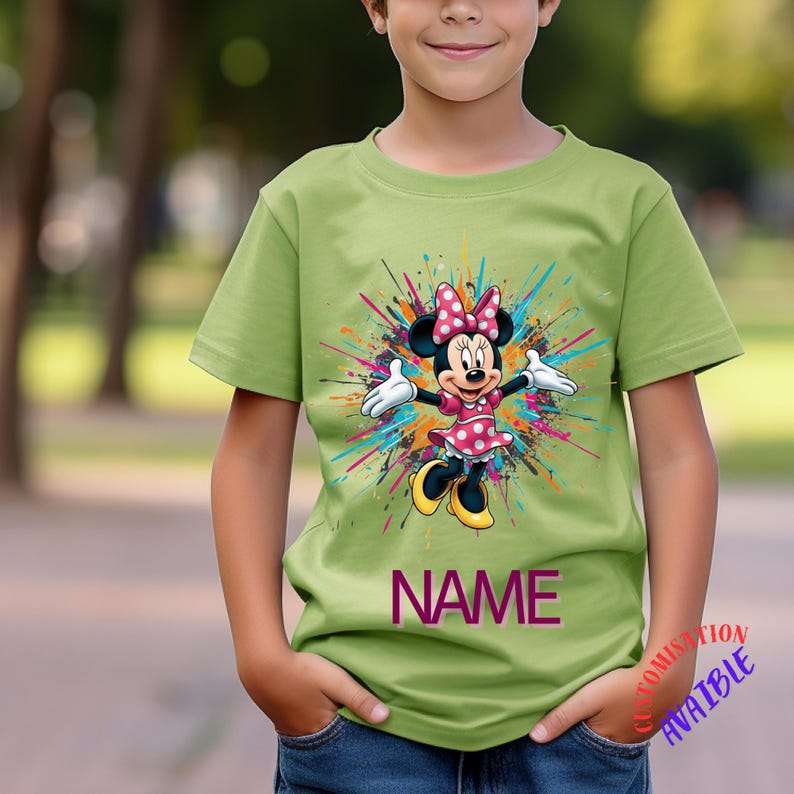 May include: A pale green t-shirt with a Minnie Mouse design. The graphic shows Minnie in a pink polka dot dress and bow, surrounded by colourful paint splashes. The word "NAME" is printed in purple.