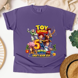 May include: A purple short-sleeve t-shirt featuring a graphic print of Toy Story characters including Woody, Buzz Lightyear, Jessie, and Bullseye. The design includes a large number 5, the text Toy Story, and the text ANDY'S ROOM 2026.