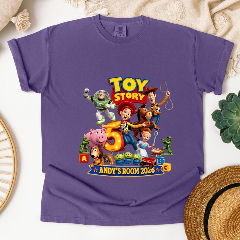 May include: A purple short-sleeve t-shirt featuring a graphic print of Toy Story characters including Woody, Buzz Lightyear, Jessie, and Bullseye. The design includes a large number 5, the text Toy Story, and the text ANDY'S ROOM 2026.