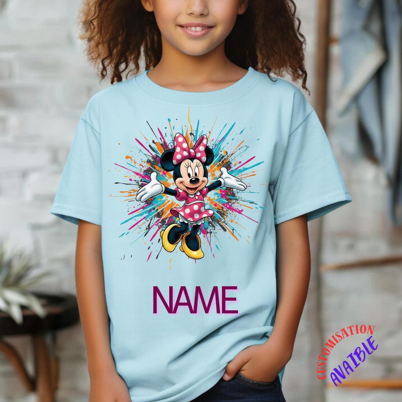 May include: Pale blue t-shirt with a Minnie Mouse graphic, wearing a pink polka dot dress and bow. The design includes colourful paint splatters and the word "NAME" in purple. A fun, personalised tee for children.