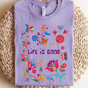 May include: A lilac-colored t-shirt with a crew neck, featuring a vibrant floral design in shades of red, blue, yellow, and orange. The words "LIFE IS GOOD" are printed in a dark purple font, centered on the shirt.