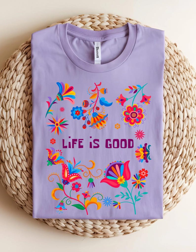 May include: A lilac-colored t-shirt with a crew neck, featuring a vibrant floral design in shades of red, blue, yellow, and orange. The words "LIFE IS GOOD" are printed in a dark purple font, centered on the shirt.