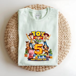 May include: A light green t-shirt featuring a Toy Story graphic with characters Woody, Buzz Lightyear, Jessie, Bullseye, Slinky Dog, Rex, and green army men surrounding a large number 5. The text reads Andy's Room 2026.
