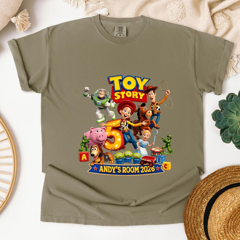 May include: A khaki short-sleeved t-shirt featuring a Toy Story graphic with characters including Buzz Lightyear, Woody, Jessie, and Bullseye. The design includes a large number 5, the text TOY STORY, and ANDY'S ROOM 2026 with various toy figures.