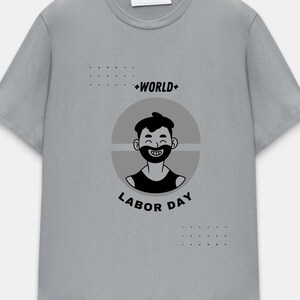 May include: A light gray t-shirt featuring a black and white graphic of a smiling person with a beard inside a gray circle. The text "+WORLD+ LABOR DAY" is above and below the graphic.