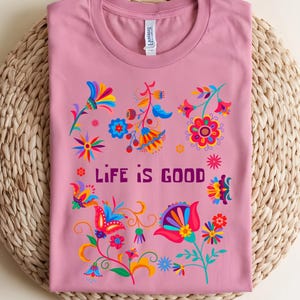 May include: A dusty rose t-shirt with a crew neck, featuring a colorful floral design and the text "LIFE IS GOOD" in purple. The flowers are in shades of red, blue, yellow, and orange. The shirt is folded on a woven mat.