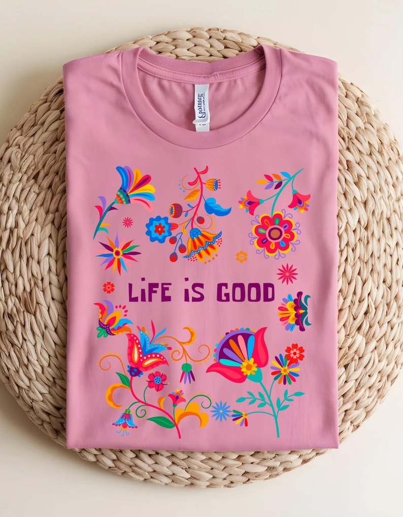 May include: A dusty rose t-shirt with a crew neck, featuring a colorful floral design and the text "LIFE IS GOOD" in purple. The flowers are in shades of red, blue, yellow, and orange. The shirt is folded on a woven mat.