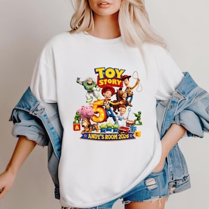 May include: A white t-shirt featuring a colorful Toy Story graphic with characters including Woody, Buzz Lightyear, Jessie, and Bullseye. The design includes a large number 5, the text Toy Story, and the text Andy's Room 2026, surrounded by toy blocks.