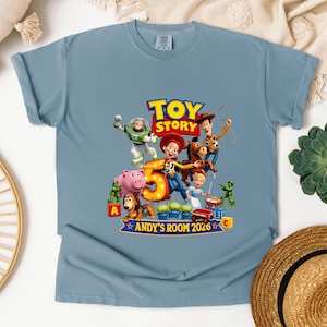 May include: A dusty blue short-sleeve t-shirt featuring a colorful graphic of Toy Story characters including Woody, Buzz Lightyear, Jessie, and Bullseye. The design includes a large number 5, the text TOY STORY, and ANDY'S ROOM 2026.