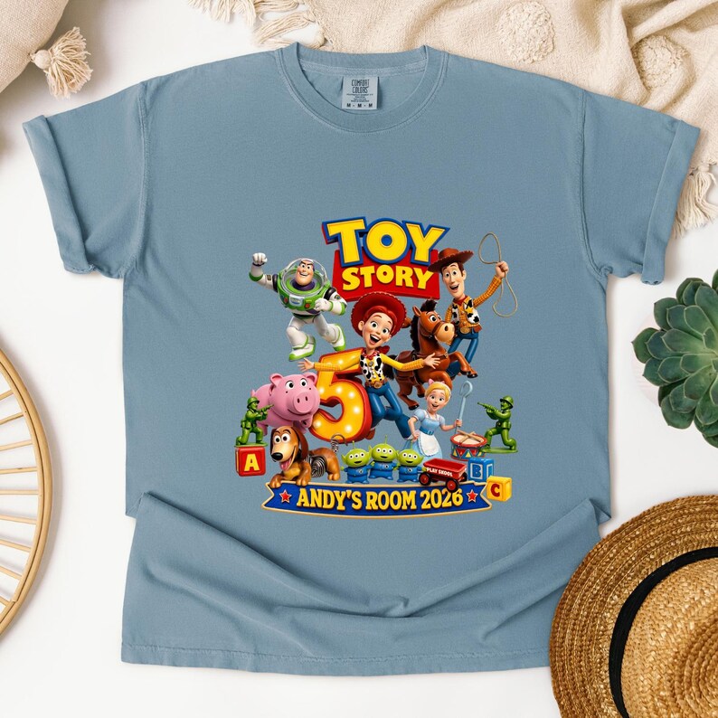 May include: A dusty blue short-sleeve t-shirt featuring a colorful graphic of Toy Story characters including Woody, Buzz Lightyear, Jessie, and Bullseye. The design includes a large number 5, the text TOY STORY, and ANDY'S ROOM 2026.