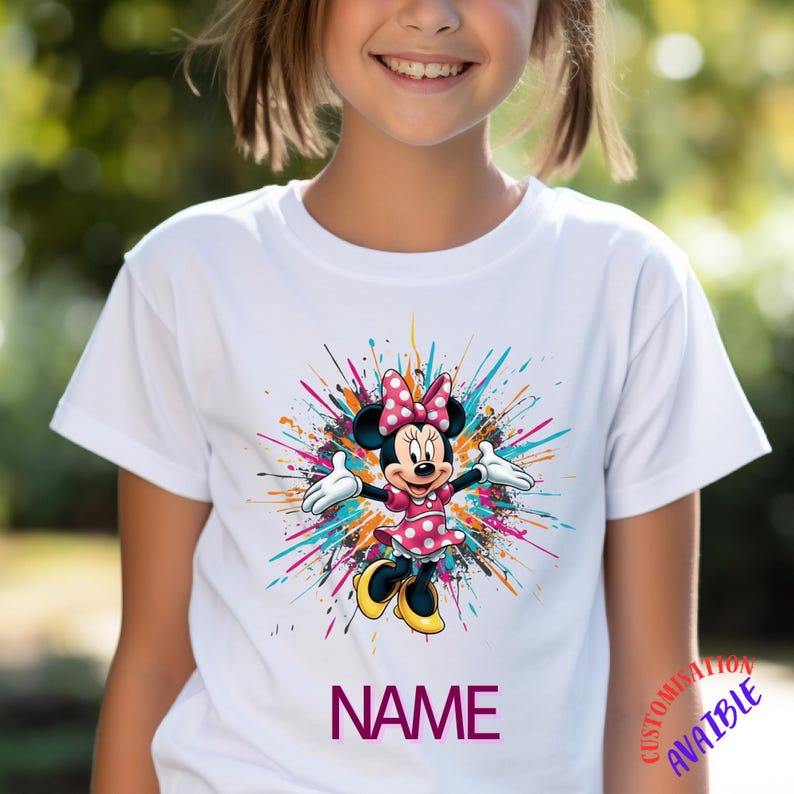 May include: White t-shirt with a Minnie Mouse graphic, wearing a pink polka dot dress and bow. The design includes colourful paint splatters and the word "NAME" in purple. A children's apparel item.
