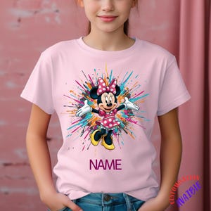 May include: A pale pink t-shirt with a Minnie Mouse graphic. The design shows Minnie Mouse in a pink polka-dot dress and bow, with a colourful paint splatter background. The word "NAME" is printed below the graphic.