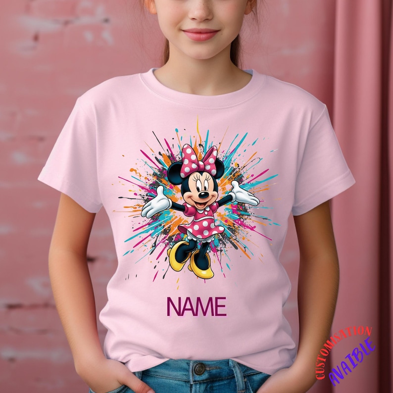 May include: A pale pink t-shirt with a Minnie Mouse graphic. The design shows Minnie Mouse in a pink polka-dot dress and bow, with a colourful paint splatter background. The word "NAME" is printed below the graphic.