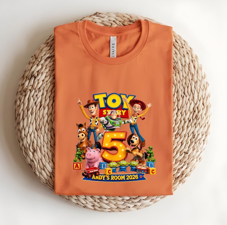 May include: An orange crew neck t-shirt featuring a Toy Story graphic with characters including Woody, Buzz Lightyear, Jessie, Bullseye, and Rex. The design includes a large number 5, alphabet blocks, and the text Andy's Room 2026.