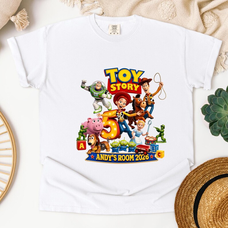 May include: A white short-sleeve t-shirt featuring a colorful Toy Story graphic with characters including Woody, Buzz Lightyear, Jessie, and Bullseye. The design includes a large number 5 and the text "TOY STORY" and "ANDY'S ROOM 2026".