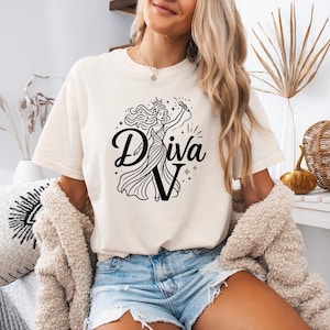 May include: A cream-colored short-sleeve t-shirt featuring a black graphic print of a person wearing a crown and holding a microphone. The text "Diva" is printed in a stylized black font, with the letter "V" integrated into the figure's dress.