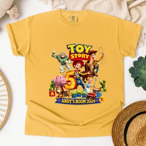 May include: A mustard yellow short-sleeve t-shirt featuring a colorful Toy Story graphic. The design includes characters like Buzz Lightyear, Woody, Jessie, and Bullseye surrounding a large number 5, with the text Toy Story and Andy's Room 2026.