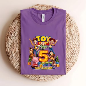 May include: A purple t-shirt featuring a graphic print of Toy Story characters including Woody, Buzz Lightyear, Jessie, Bullseye, Hamm, Slinky Dog, and green army men surrounding a large yellow number 5 with the text Andy's Room 2026.