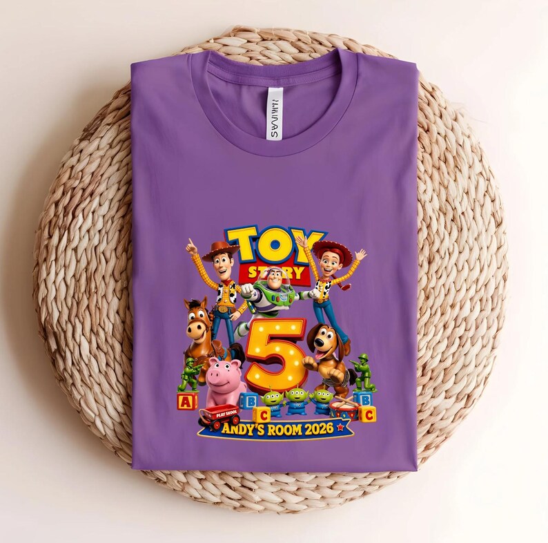 May include: A purple t-shirt featuring a graphic print of Toy Story characters including Woody, Buzz Lightyear, Jessie, Bullseye, Hamm, Slinky Dog, and green army men surrounding a large yellow number 5 with the text Andy's Room 2026.
