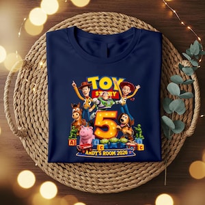 May include: A navy blue t-shirt featuring a graphic print of Toy Story characters including Woody, Buzz Lightyear, Jessie, Bullseye, Slinky Dog, and green soldiers. A large orange number 5 is centred with the text ANDY'S ROOM 2026 below it.