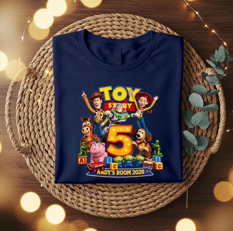May include: A navy blue t-shirt featuring a graphic print of Toy Story characters including Woody, Buzz Lightyear, Jessie, Bullseye, Slinky Dog, and green soldiers. A large orange number 5 is centred with the text ANDY'S ROOM 2026 below it.